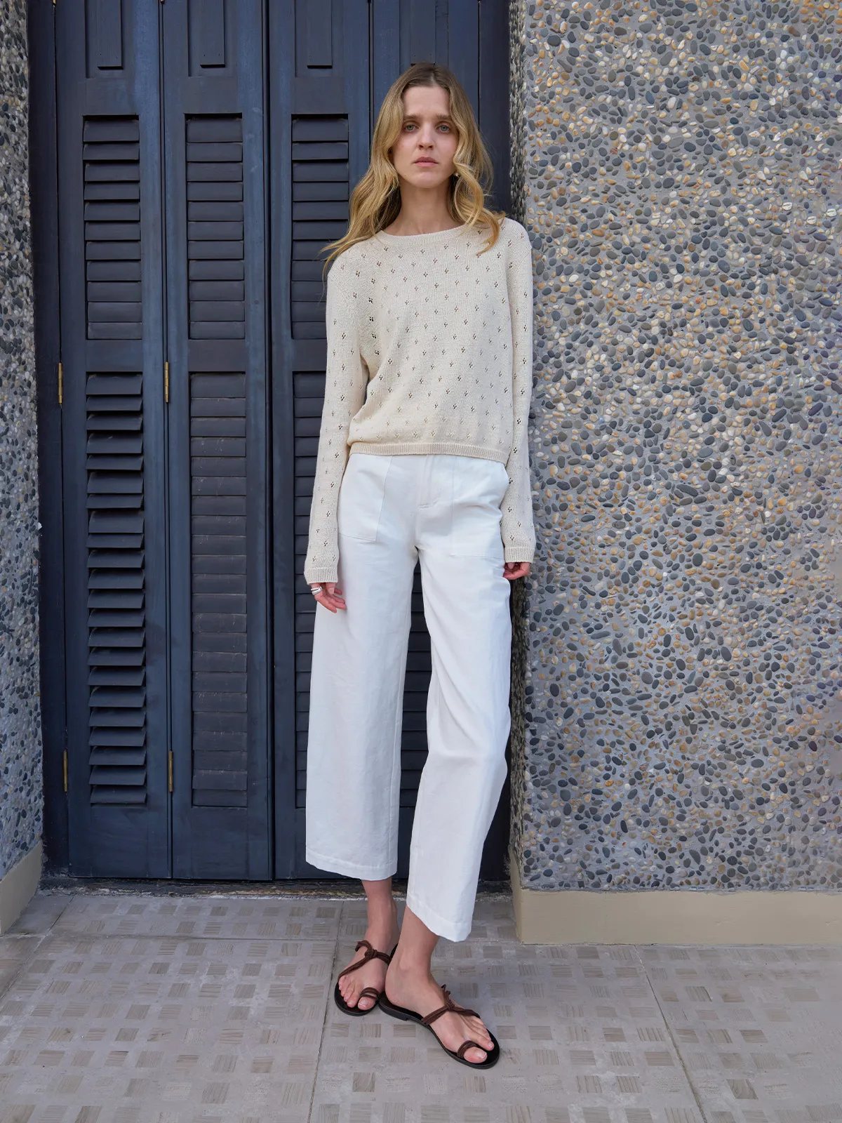 Model wearing KIANA openwork knit top with ivory wide-leg linen trousers against cobalt shutter and pebble wall