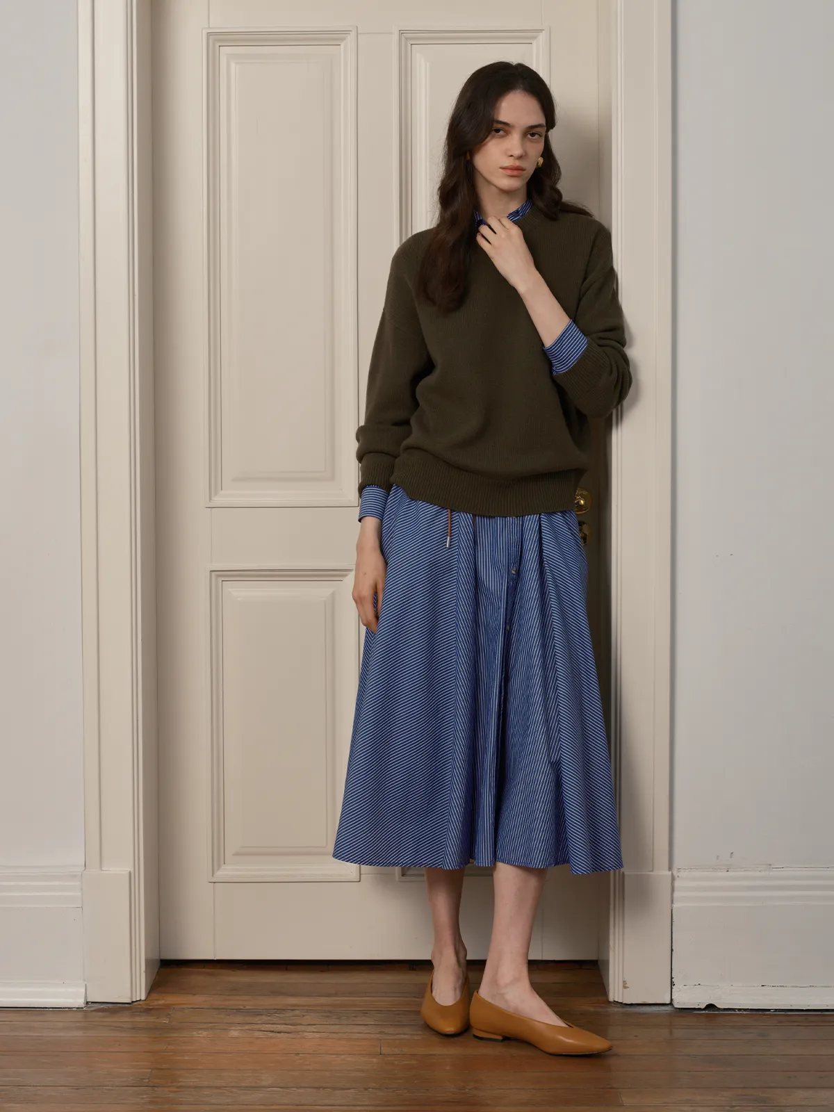Side profile of model wearing blue stripe midi skirt with olive sweater, hands in pockets, against white door
