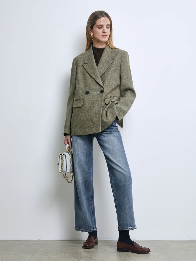 Side view of a model wearing a Sage Green pure wool tweed double-breasted blazer with notched lapels and flap pockets, paired with jeans and holding a small bag.
