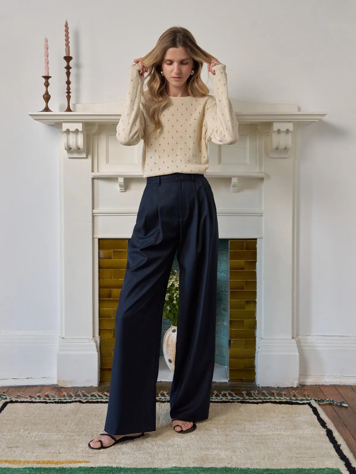 Full-length view of model in cream knit sweater and ROGAN wide leg trousers, standing before fireplace