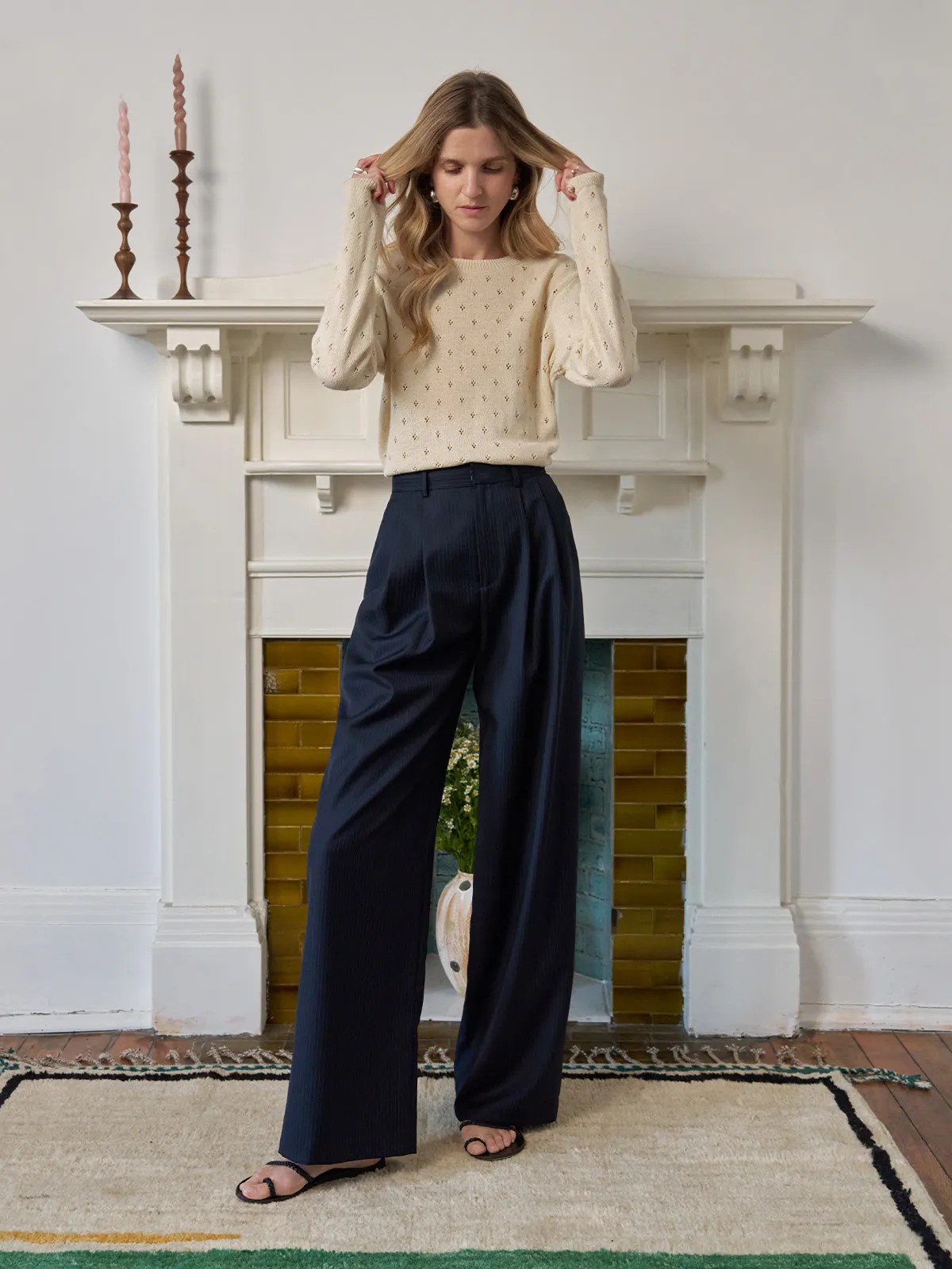 Full-length view of model in cream knit sweater and ROGAN wide leg trousers, standing before fireplace
