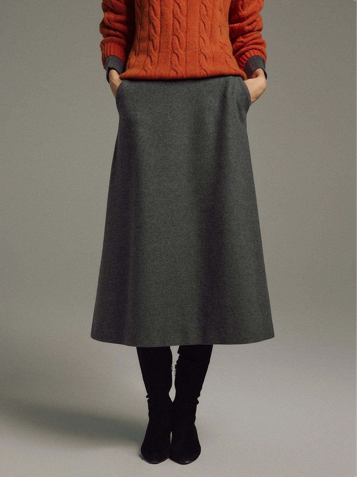 Front view of a model in a Gray wool-blend A-line midi skirt, cable-knit sweater, and boots, hands on hips.