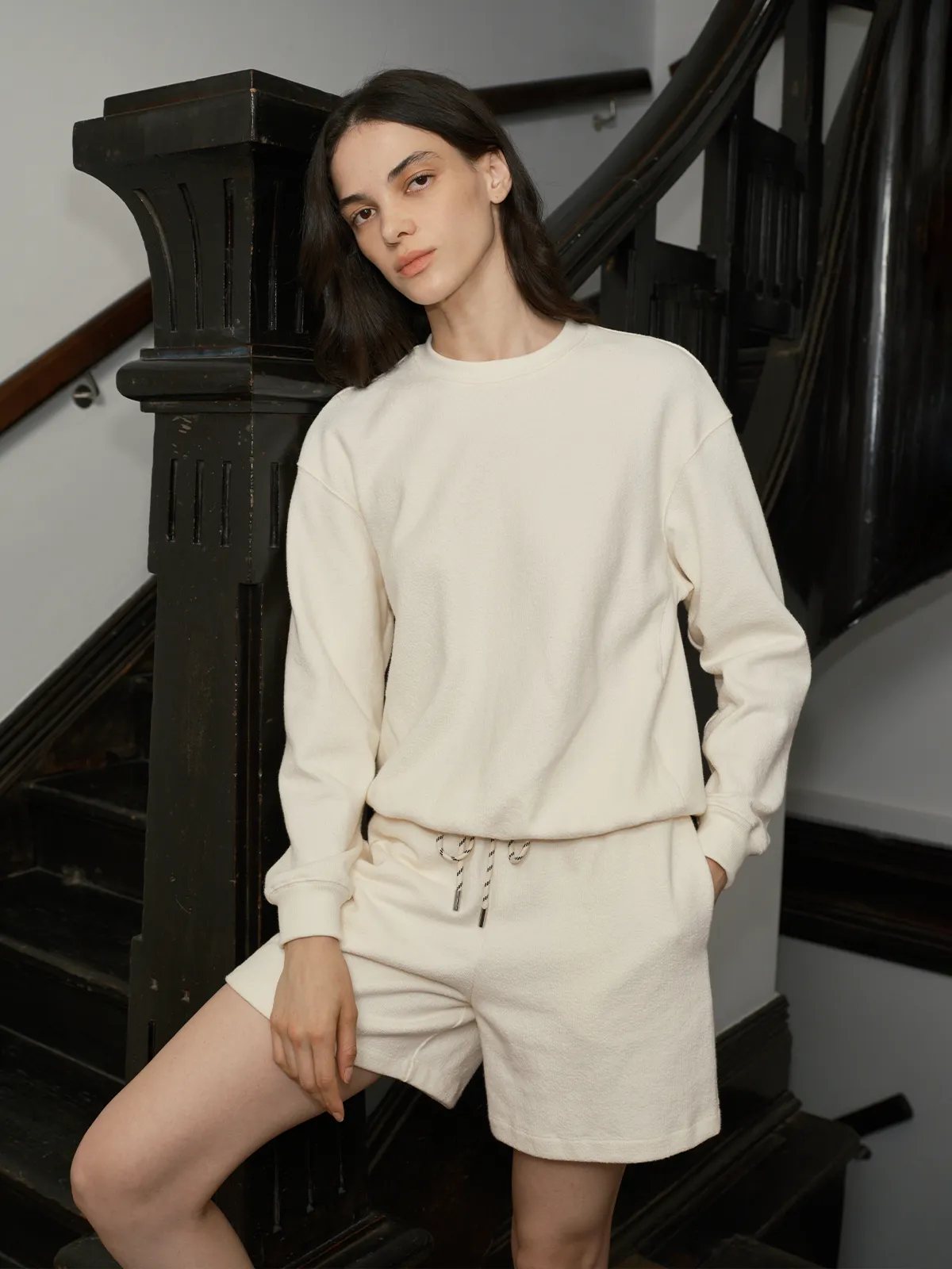 Model in ivory SOREN sweatshirt and shorts posing beside black staircase newel post