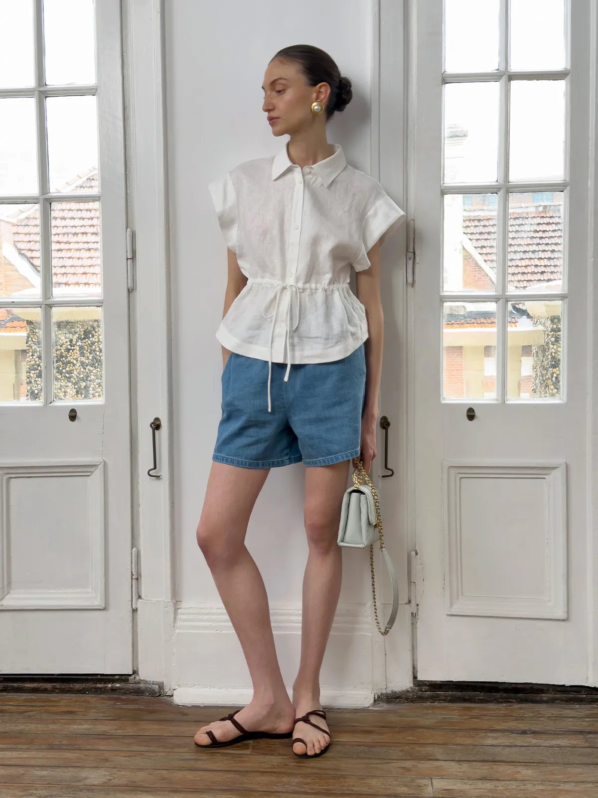 Model leaning against white door wearing MAREN linen shirt with denim shorts, chain-strap bag, and flat sandals