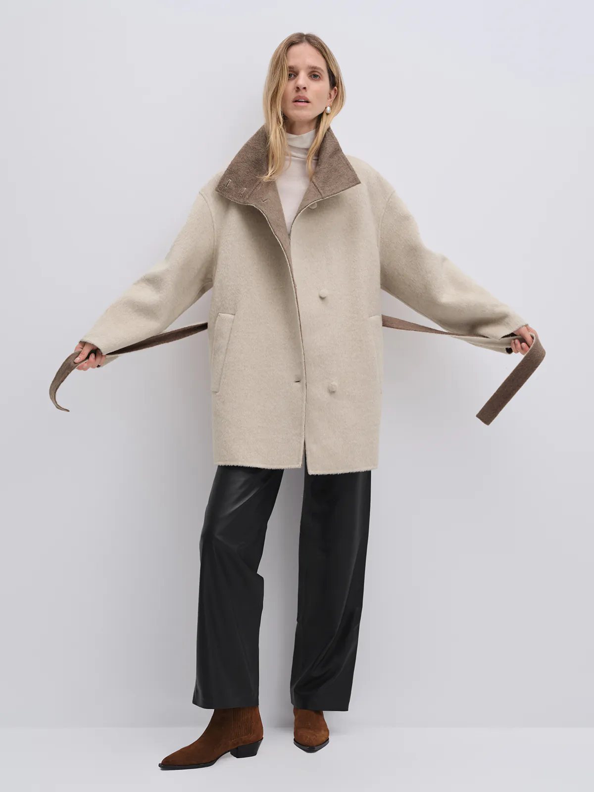 Model holding the untied belt of the Oatmeal reversible two-tone long coat, arms extended to show sleeve drape.