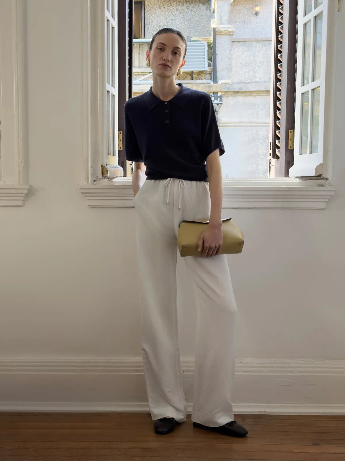 Model in navy polo and ivory linen wide-leg trousers holding mustard clutch by open window