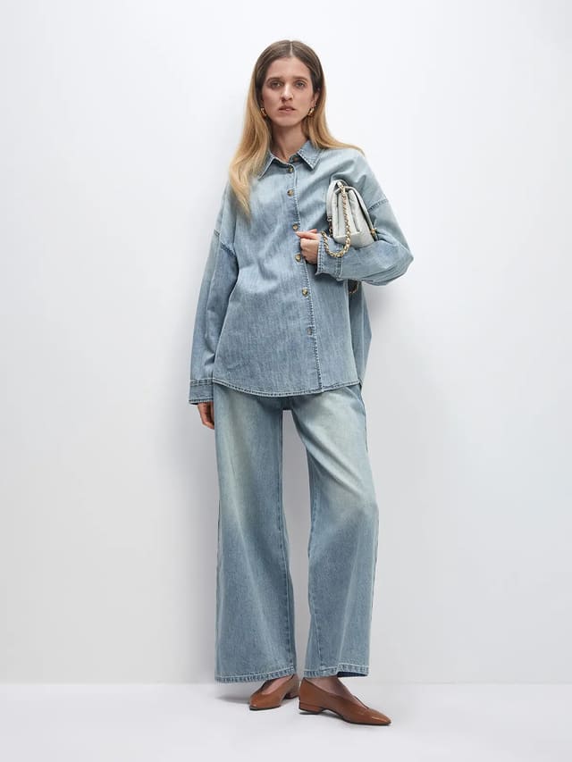 Model wears a light blue oversized denim shirt with button front and relaxed fit, paired with matching wide-leg jeans.
