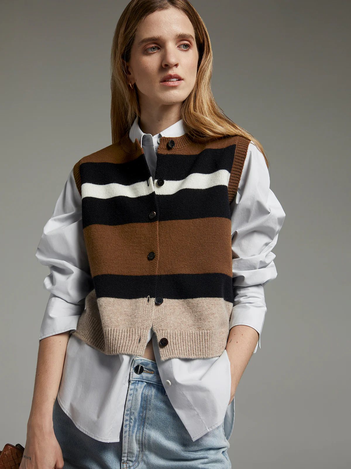 Model seated wearing a sleeveless striped patchwork knit vest in brown with contrasting bands paired with pants.