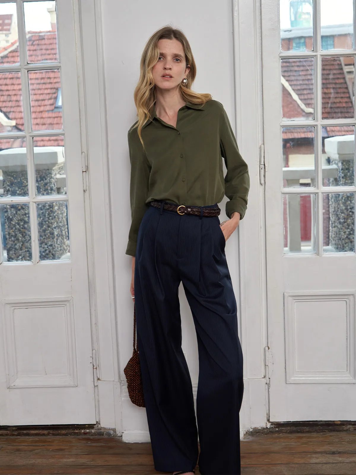 Model in olive shirt and navy pleated trousers wearing PLAIT belt near French doors