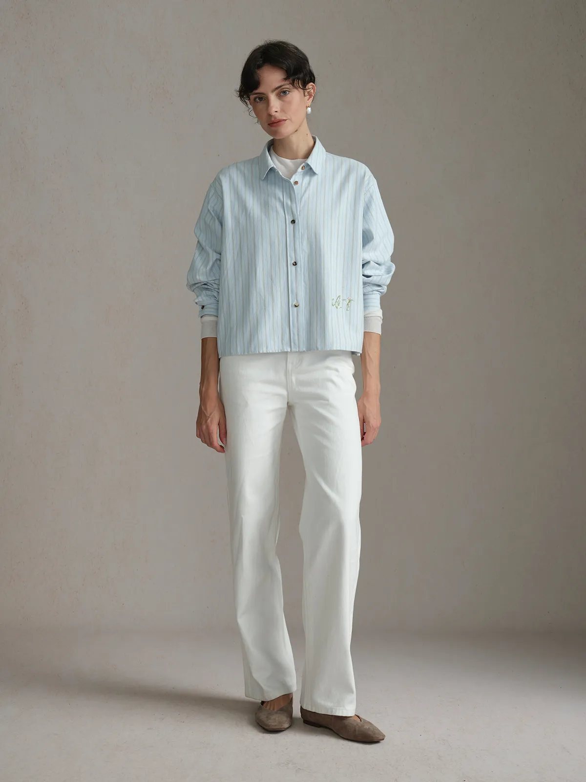 Full-body front view of model wearing PIXIE shirt with white trousers and pearl earrings, minimalist studio setting