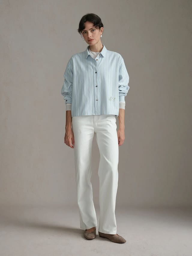 Full-body front view of model wearing PIXIE shirt with white trousers and pearl earrings, minimalist studio setting