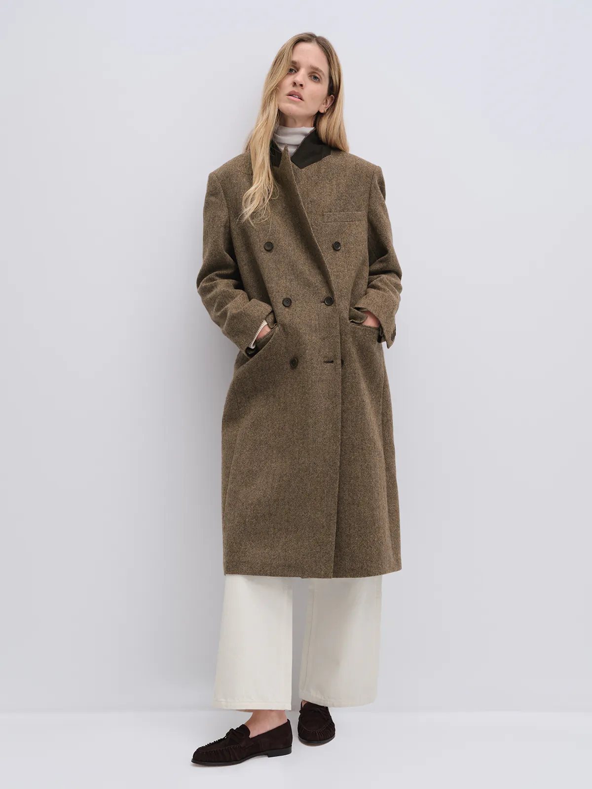 Front-facing model wearing a brown wool and alpaca tweed long coat over a plaid shirt and wide-leg jeans.