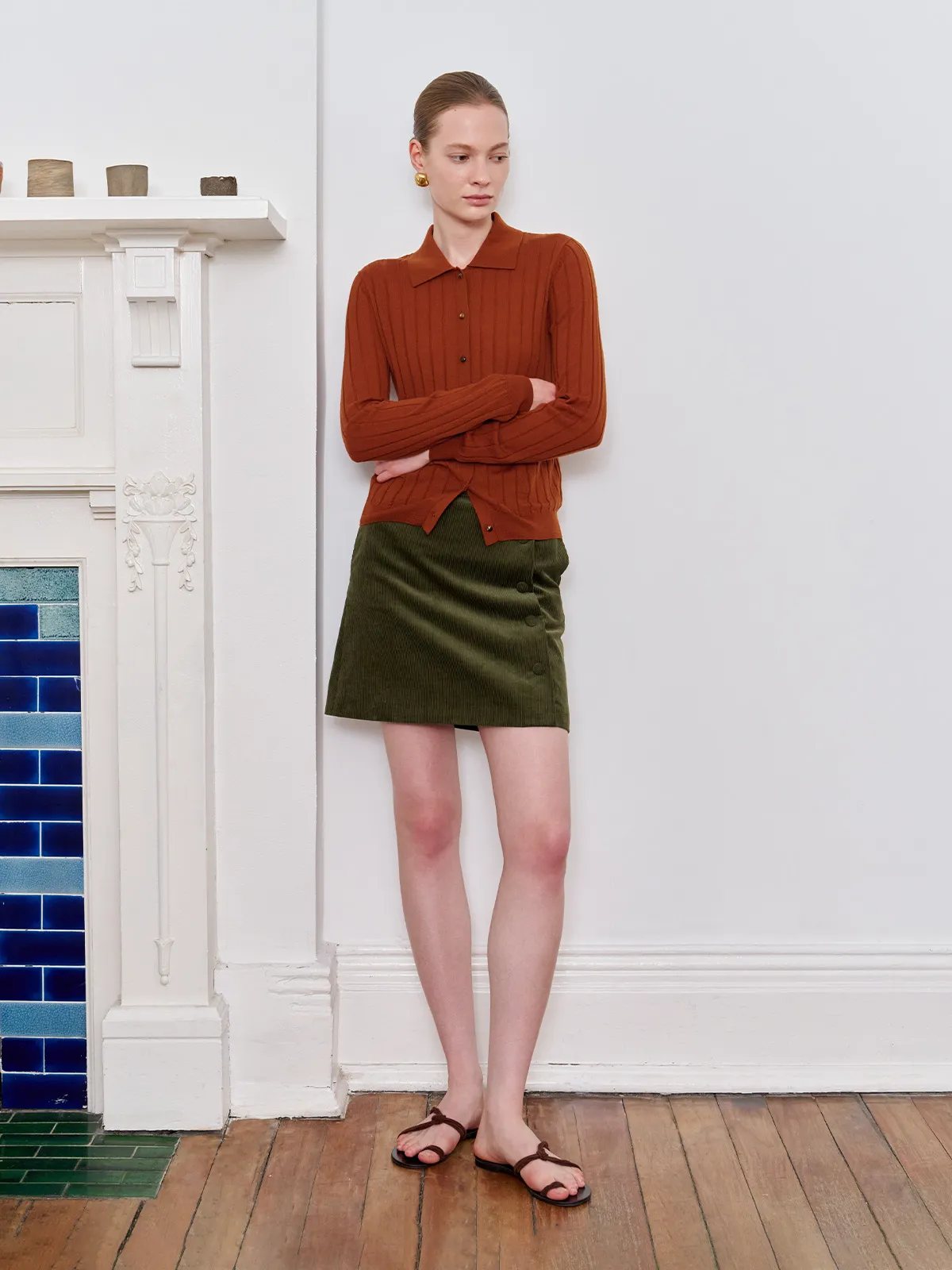 Model leaning against white fireplace with blue tile inset, wearing YUKI skirt and rust knit