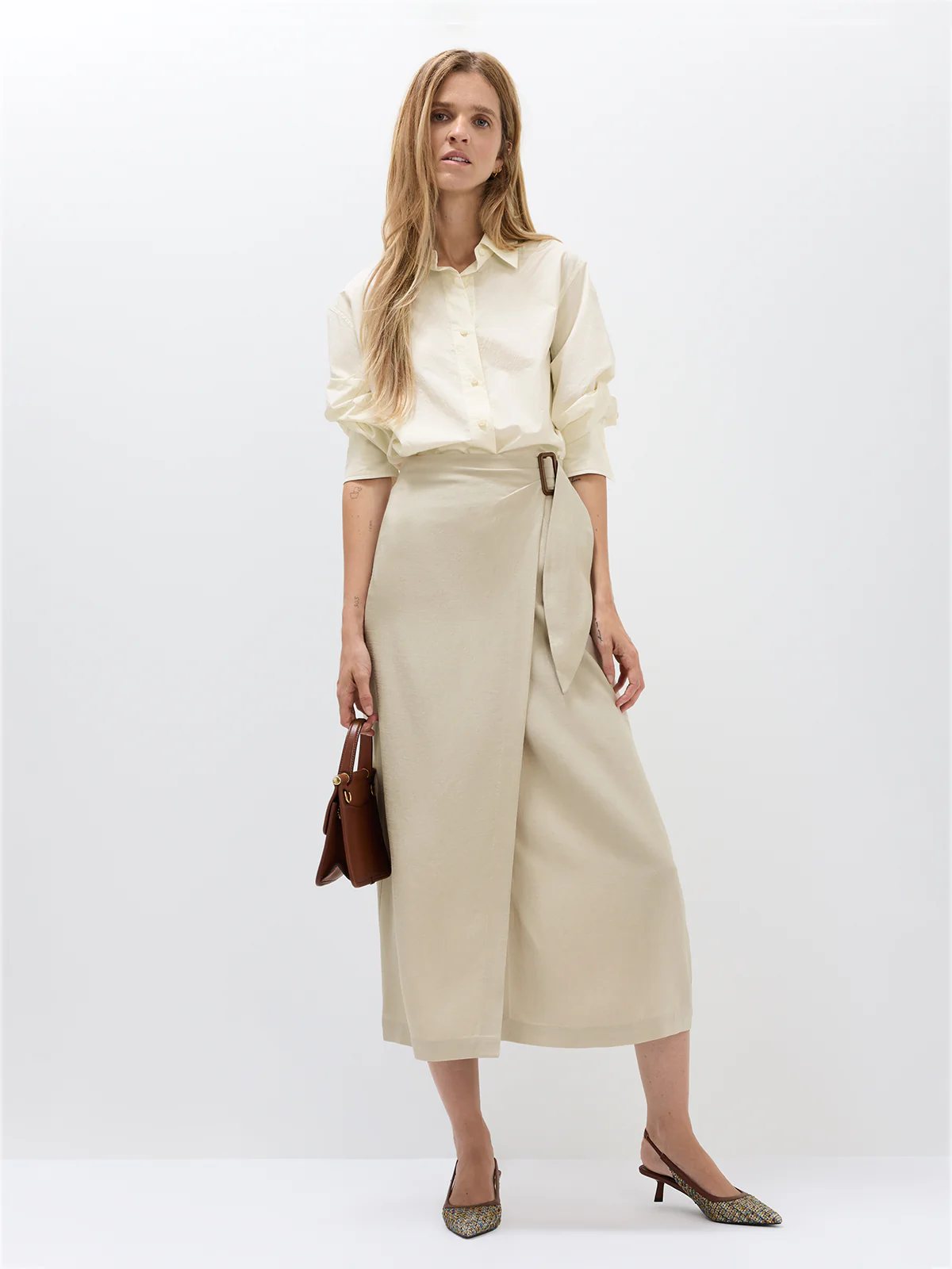 Side view of model in the Oatmeal acetate wrap midi skirt with a top, clutch, and flats.