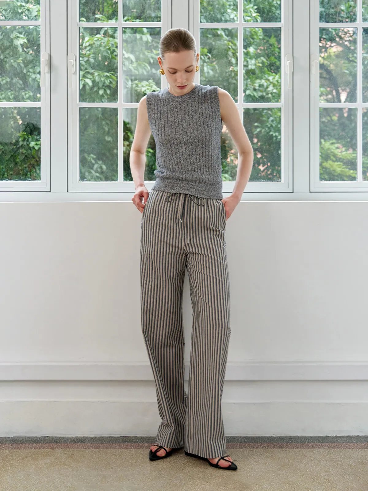 Model standing by window wearing WHIMSY trousers with grey ribbed tank, hands in pockets, natural light