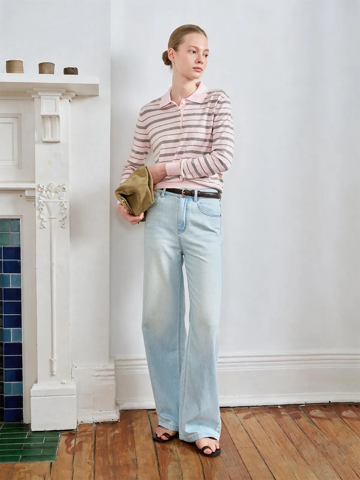 Side profile of model in HELENA striped polo top, light wash jeans, and brown sandals beside fireplace
