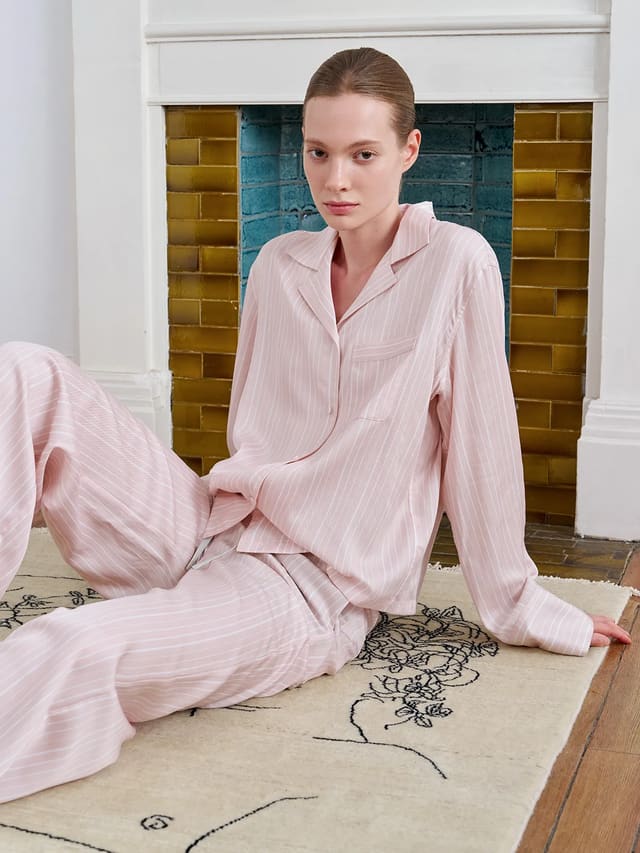 Full LARISSA pink stripe pajama set worn by model seated before vintage fireplace with gold and teal tiles