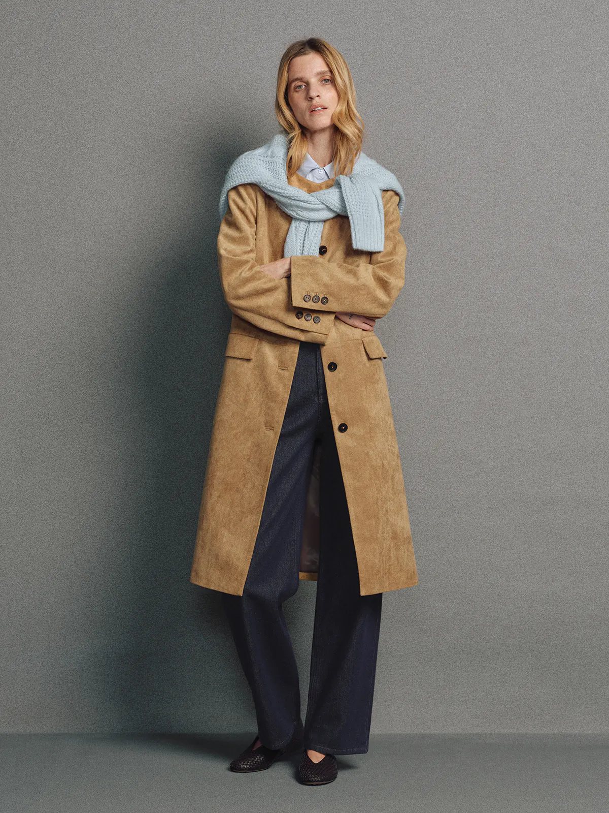 Model wearing a camel faux suede collarless long coat with front buttons, styled with a light scarf and dark trousers.