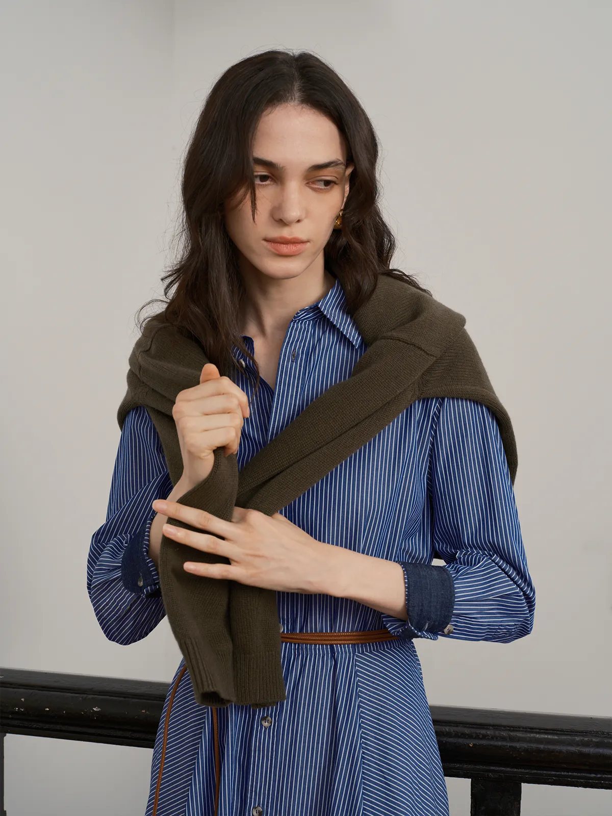 Model adjusting olive green knit vest over blue stripe shirt dress, highlighting layering versatility