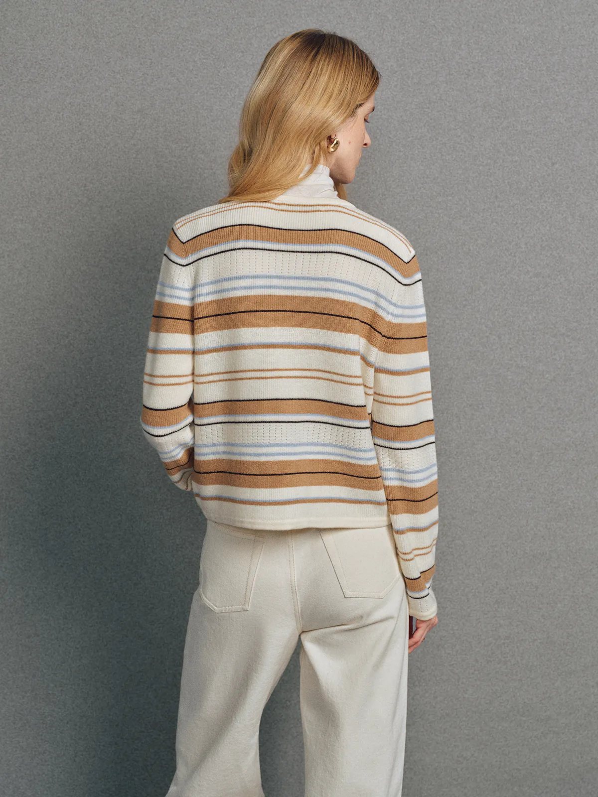Back view of the Camel multicolour stripe V-neck cardigan worn over a turtleneck with light trousers.
