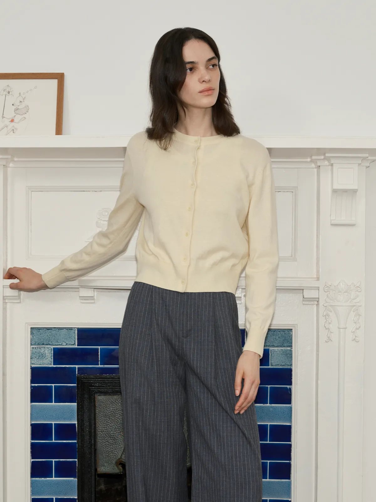 Model in BINX cardigan at white fireplace with blue tile insert, hands resting on mantel