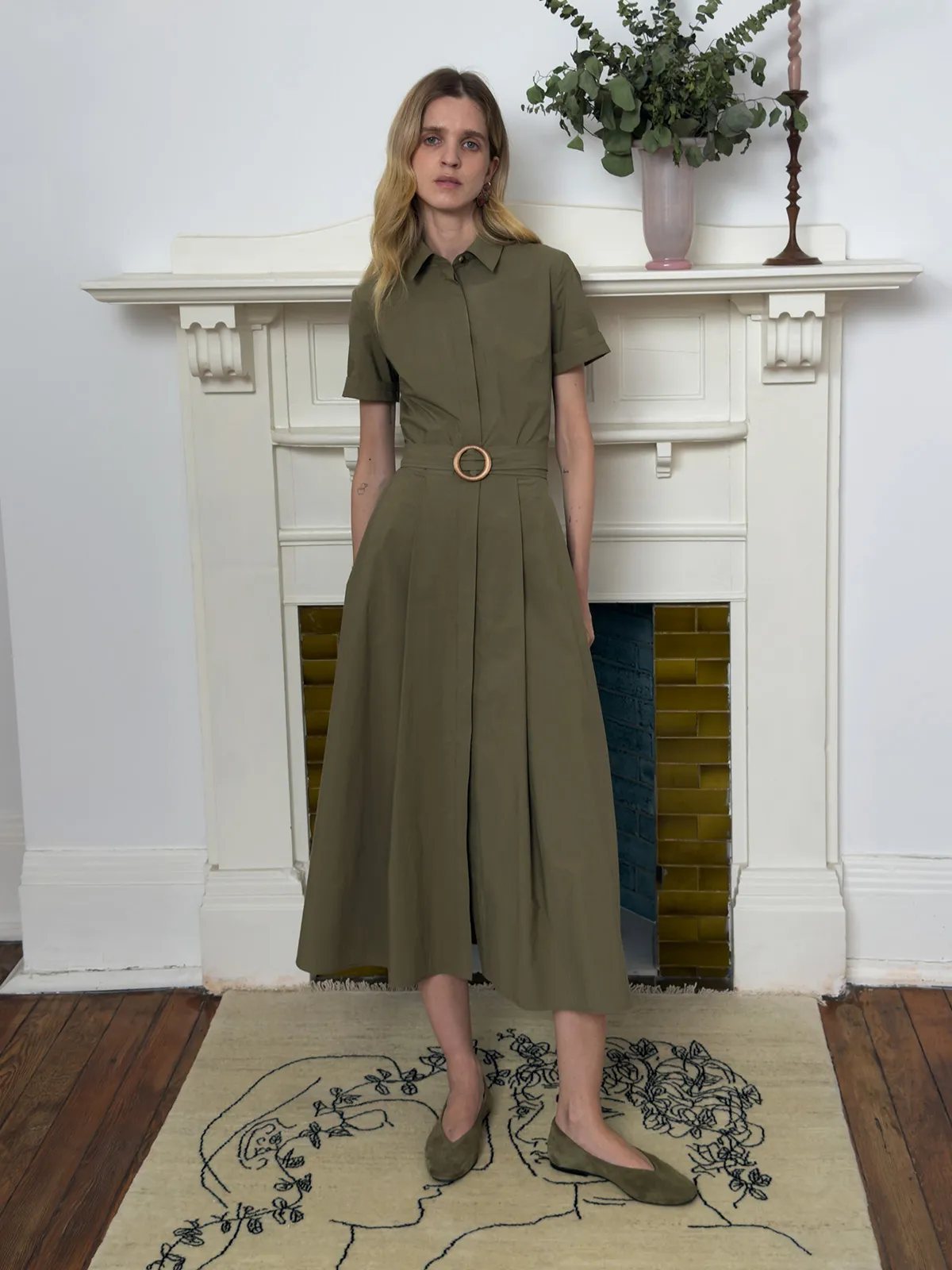 Andreatang IMANI olive green short sleeve shirt dress with wooden ring belt, model standing front view on embroidered rug