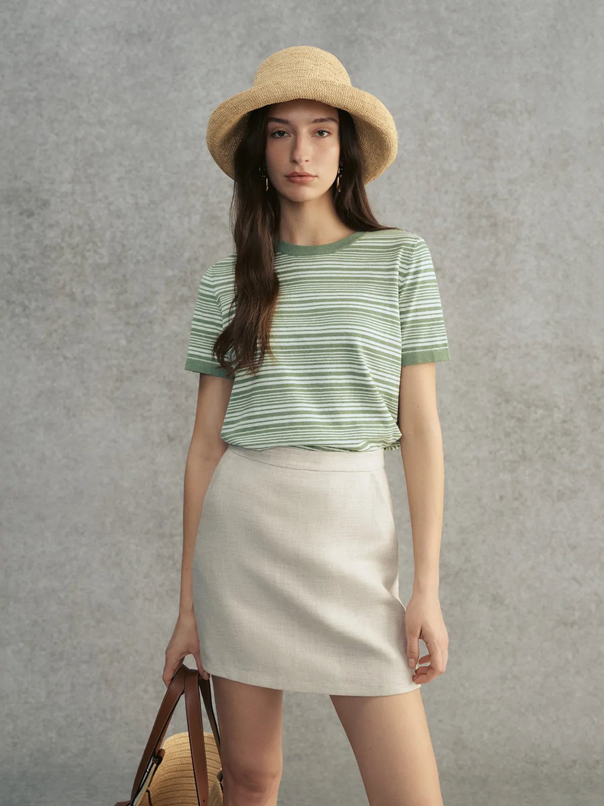 Model in sage green striped JUSTINE t-shirt, cream linen mini skirt, and wide-brimmed straw hat holding leather bag