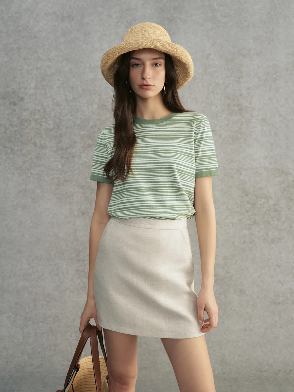 Model in sage green striped JUSTINE t-shirt, cream linen mini skirt, and wide-brimmed straw hat holding leather bag
