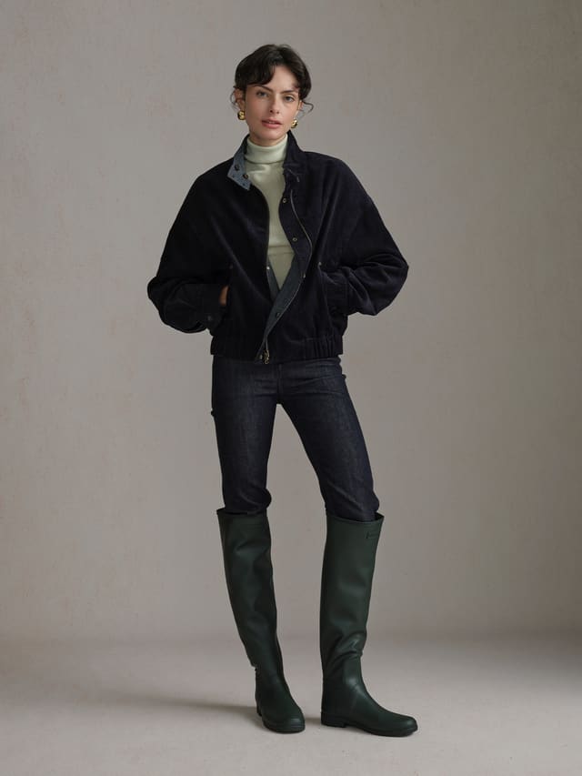 Model wearing the Navy Andreatang CHARLIE jacket, corduroy side out, with a light high-neck top, dark jeans and tall green boots.