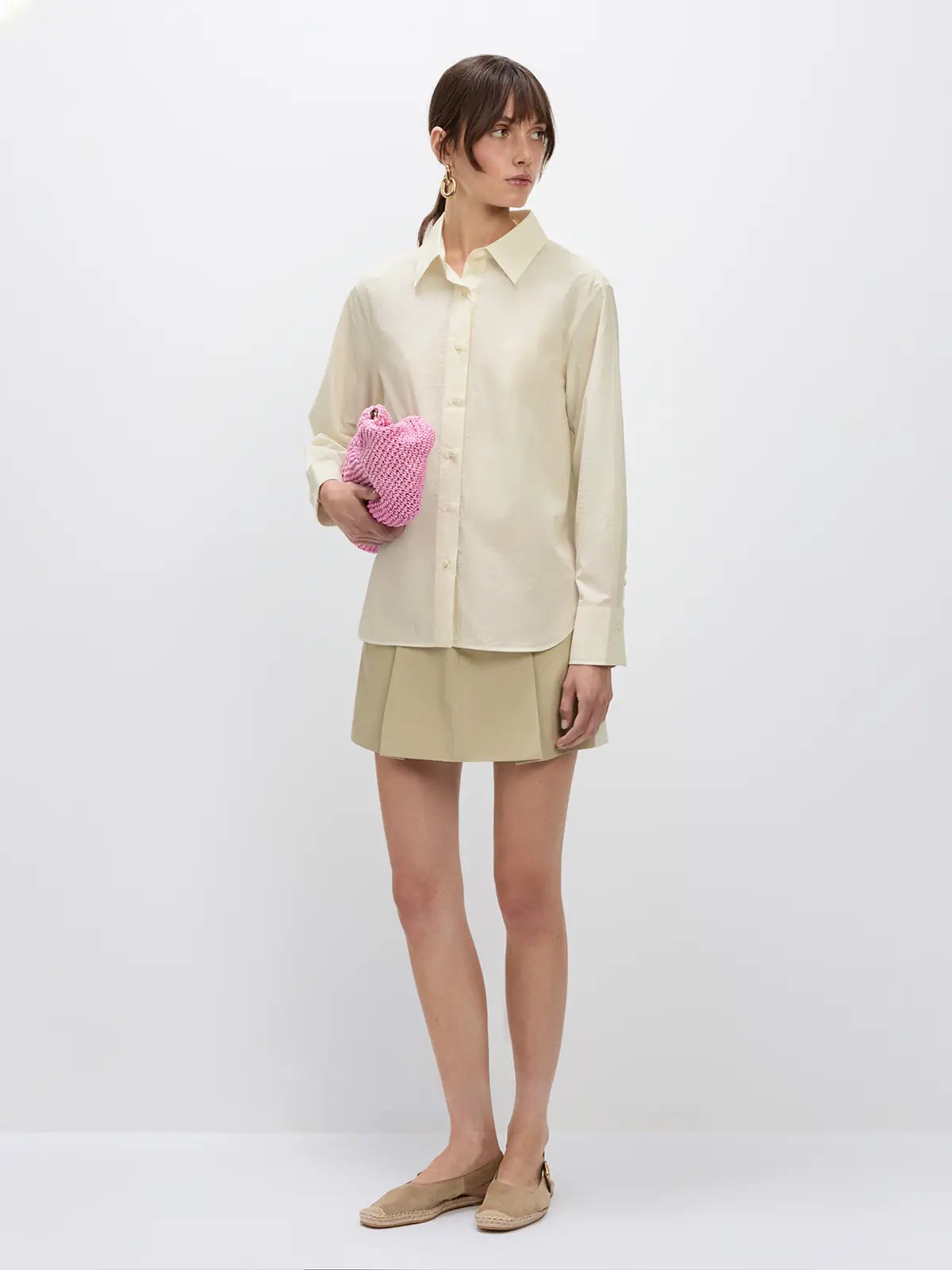 Model holding a clutch, wearing the Beige NAOMI shirt tucked into a beige pleated mini skirt.