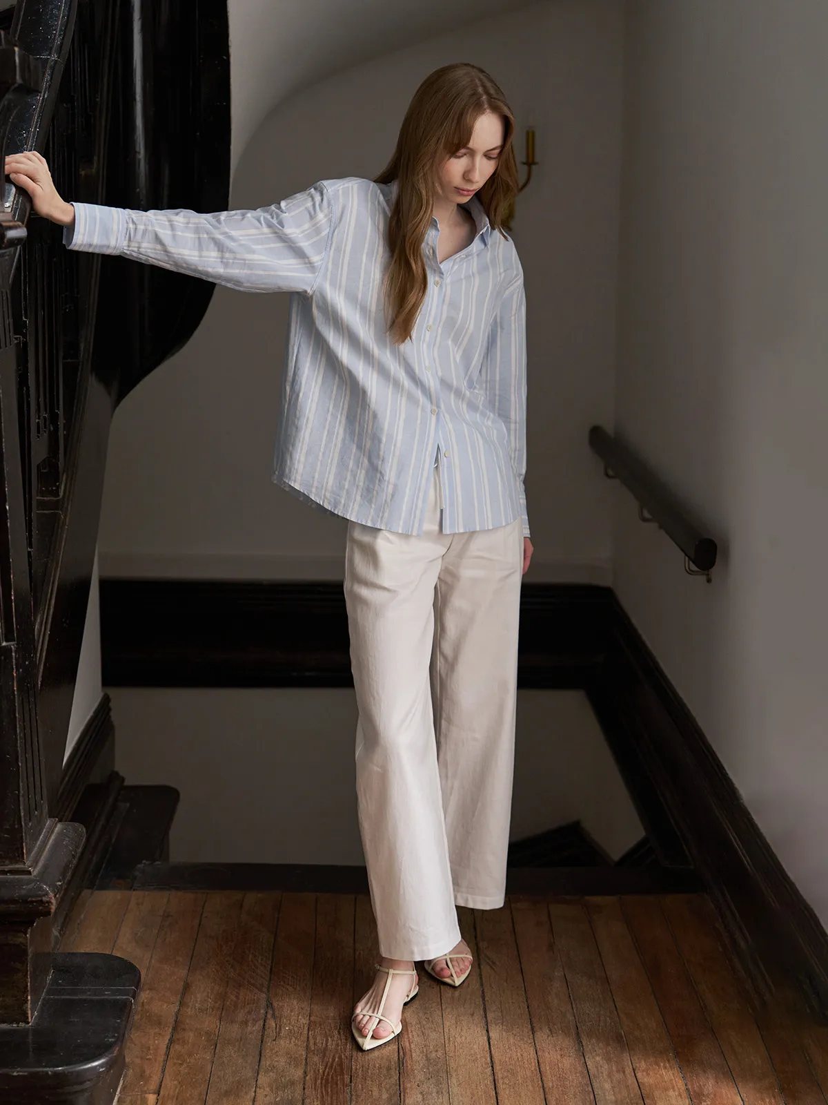 Model on wooden staircase wearing white LEIGHTON trousers with striped shirt, showing back drape and movement