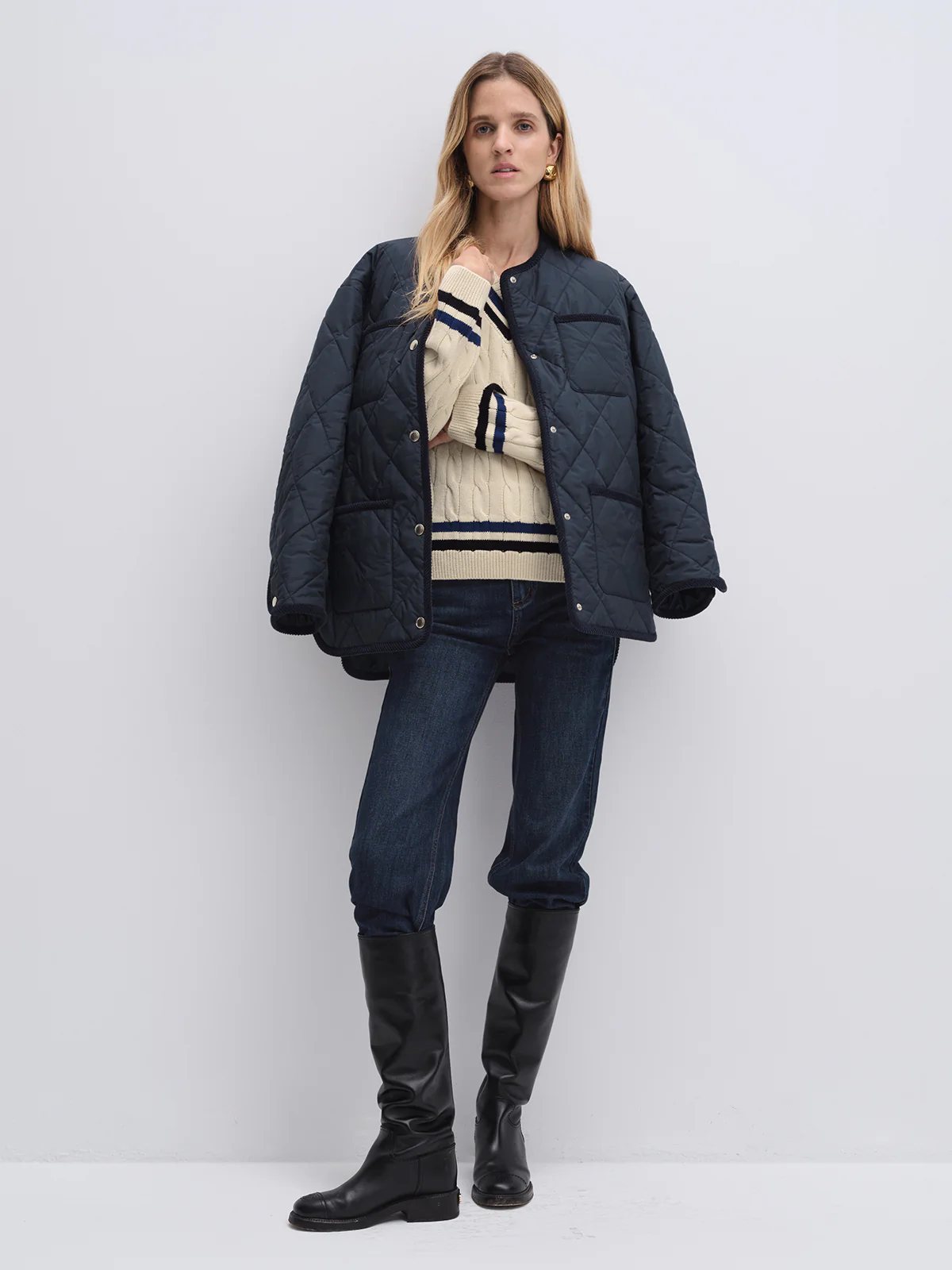 Model wearing navy quilted textured jacket open over striped sweater, paired with dark jeans and knee-high boots.