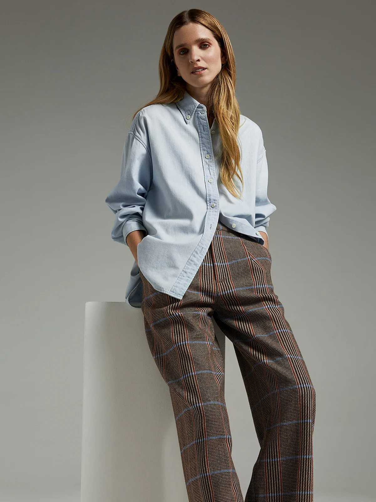 Model wearing brown wool British check trousers and matching blazer with light shirt, cap, and holding a brown handbag.