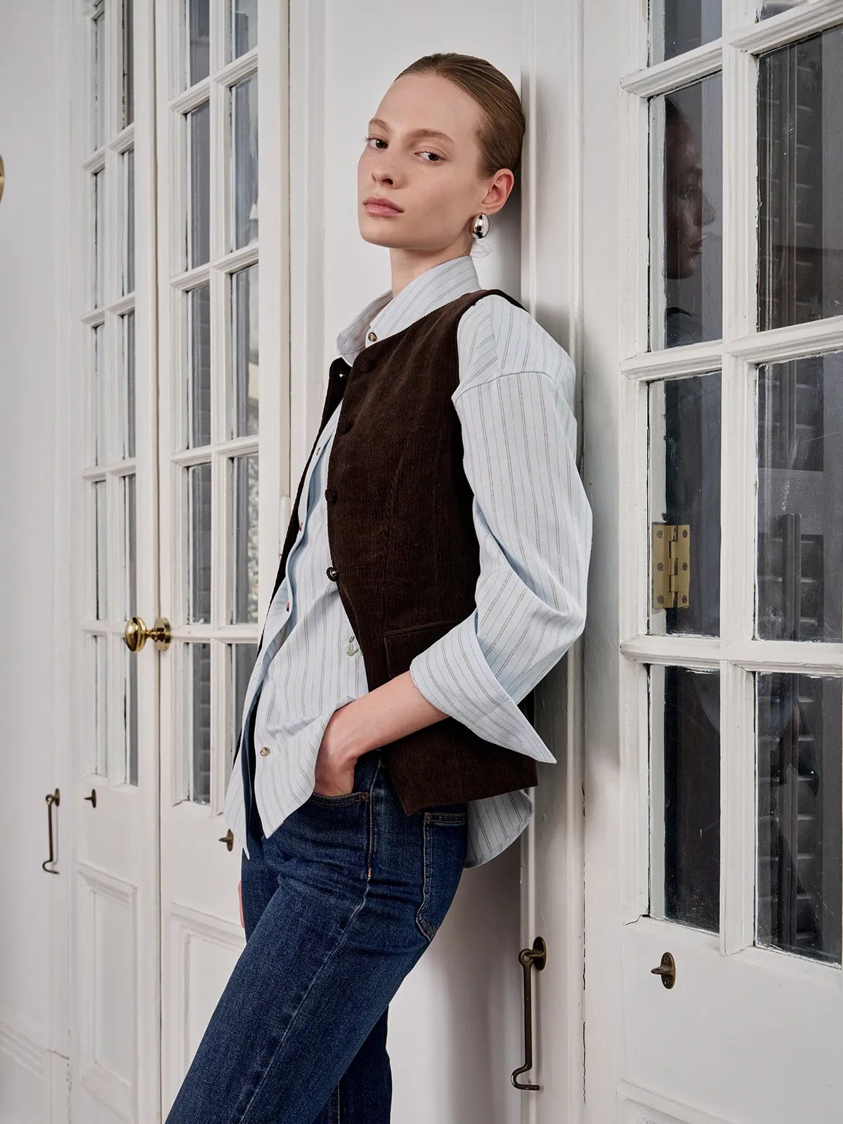 Side profile of model in TILLY shirt layered under brown corduroy vest, leaning against white French doors