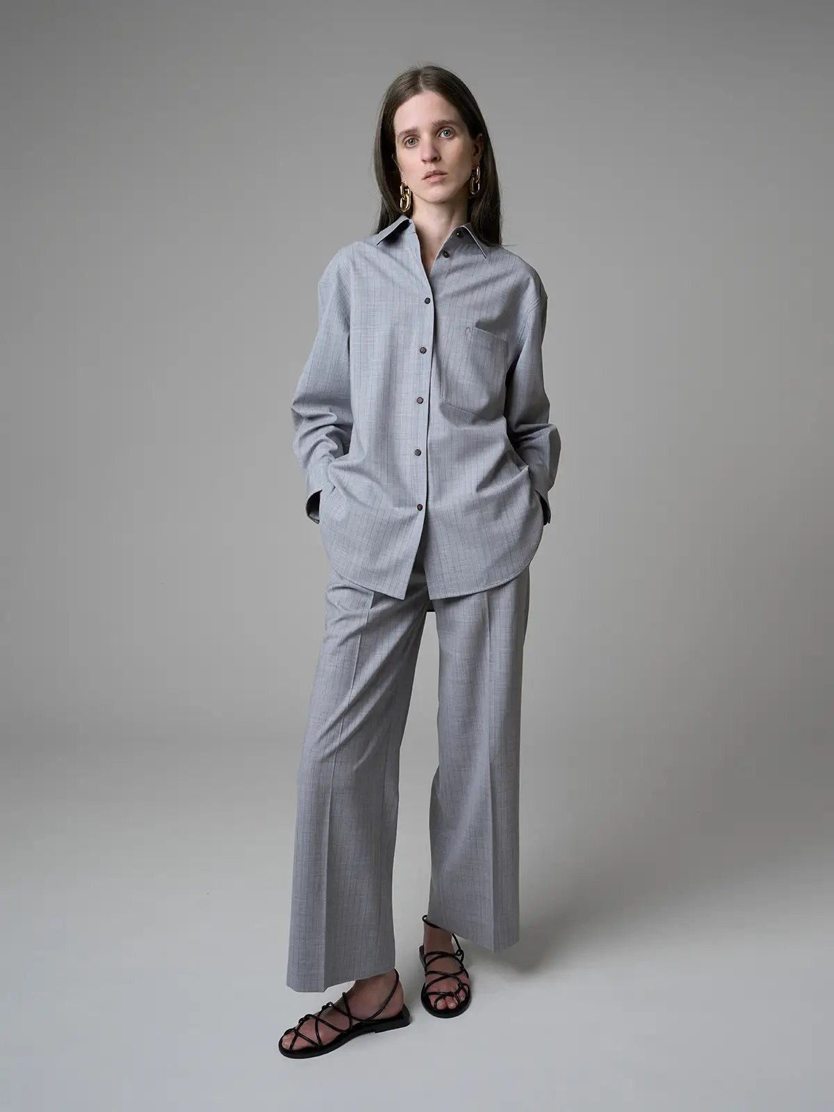 Model wearing a loose grey striped wool shirt with full-length sleeves and matching trousers, hands in pockets, front view.