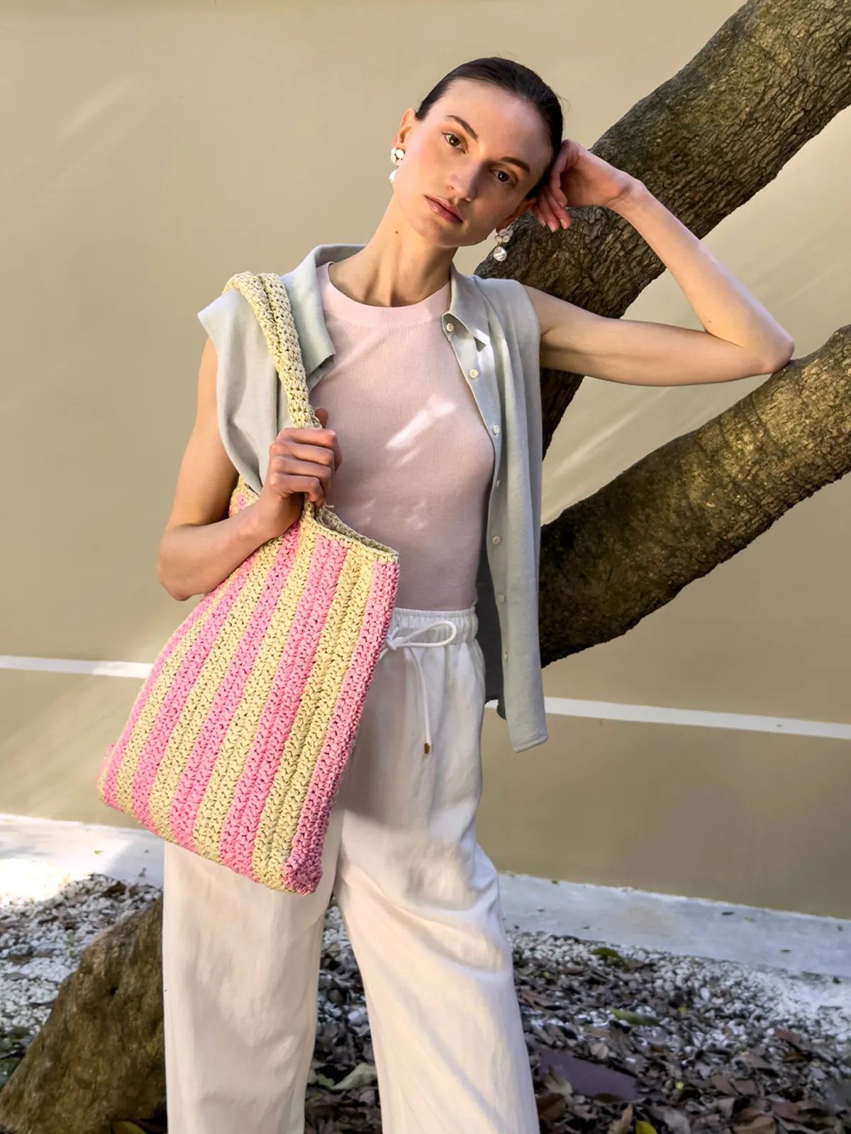 Model outdoors leaning against tree wearing MARGOT knit shirt over pink knit top with white drawstring pants and striped tote