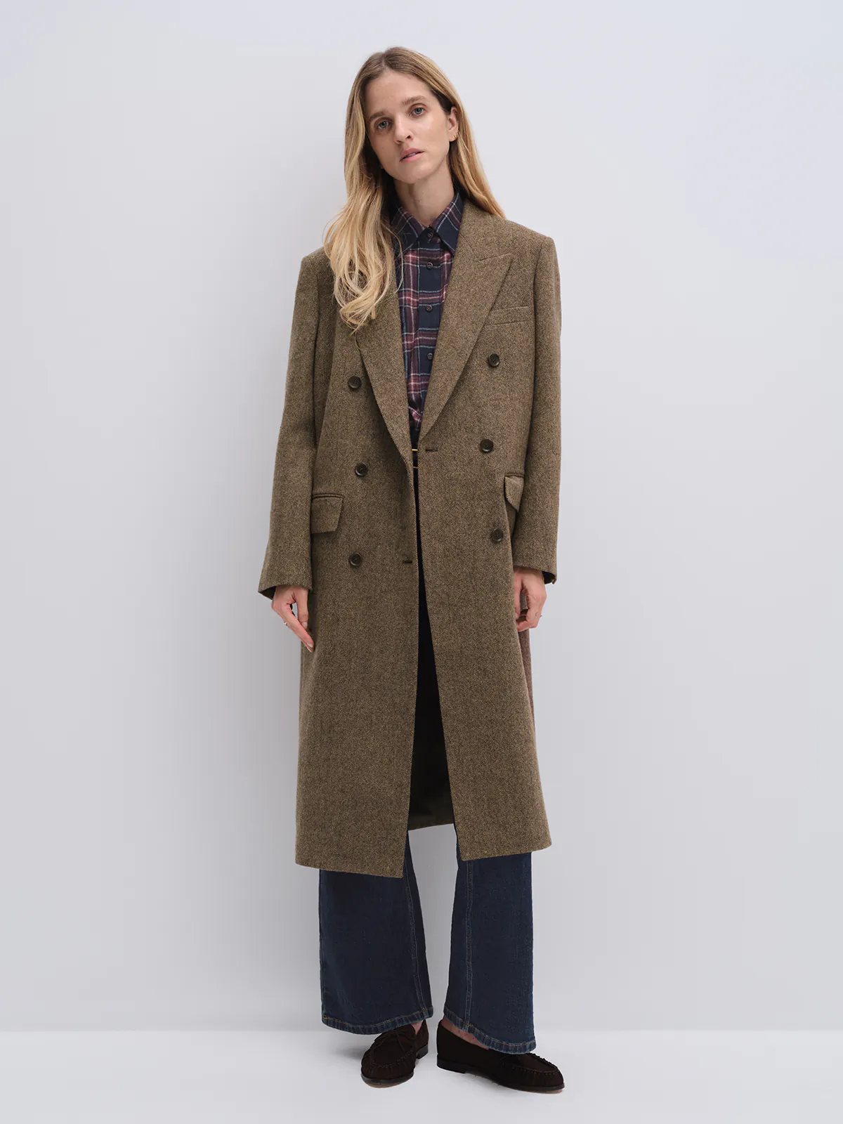 Model in a brown wool and alpaca tweed long coat holding a woven clutch, paired with trousers and boots.