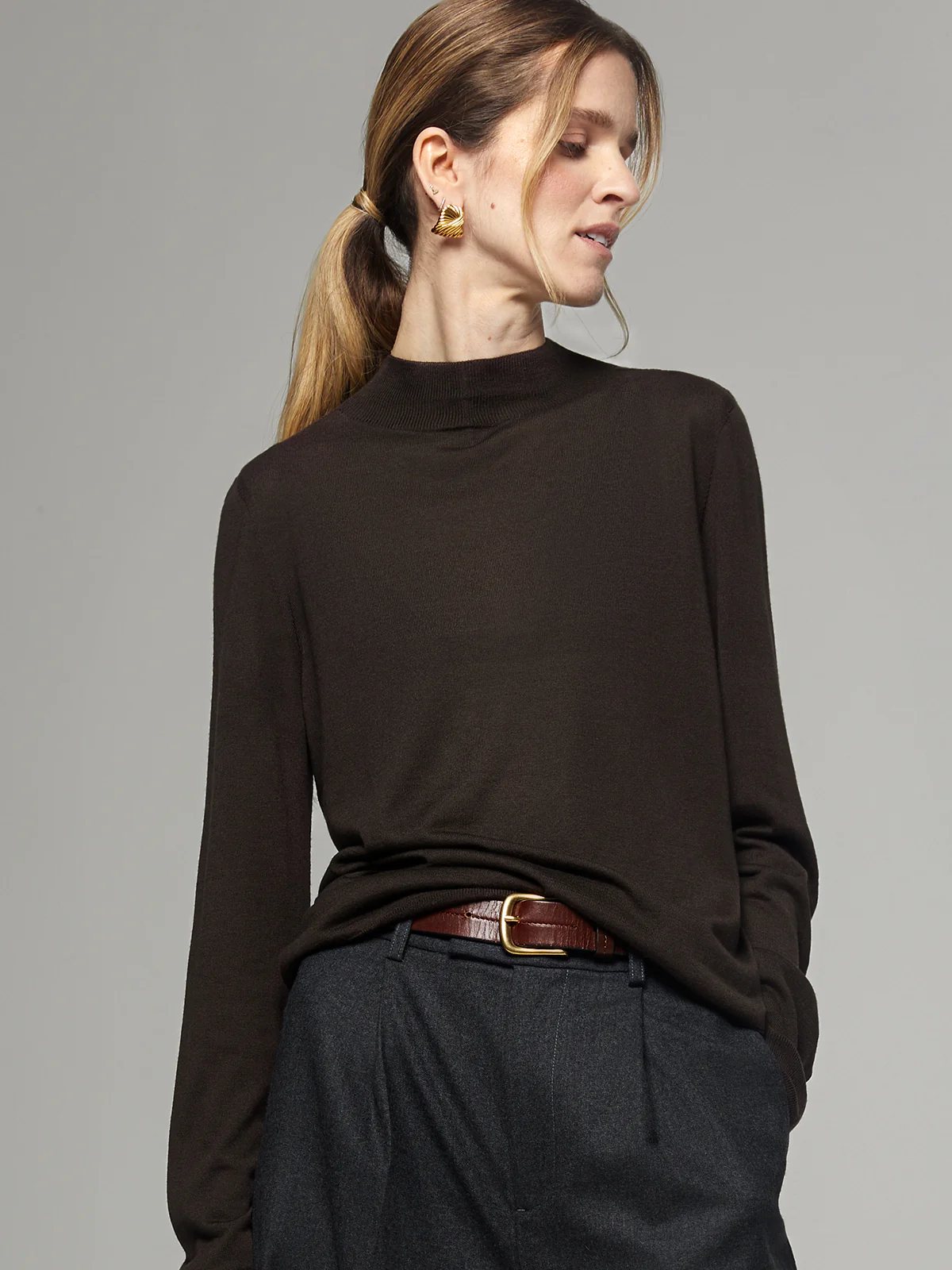 Model in a black silk lyocell mock-neck top with long sleeves, hands in pockets of trousers, wearing hoop earrings.