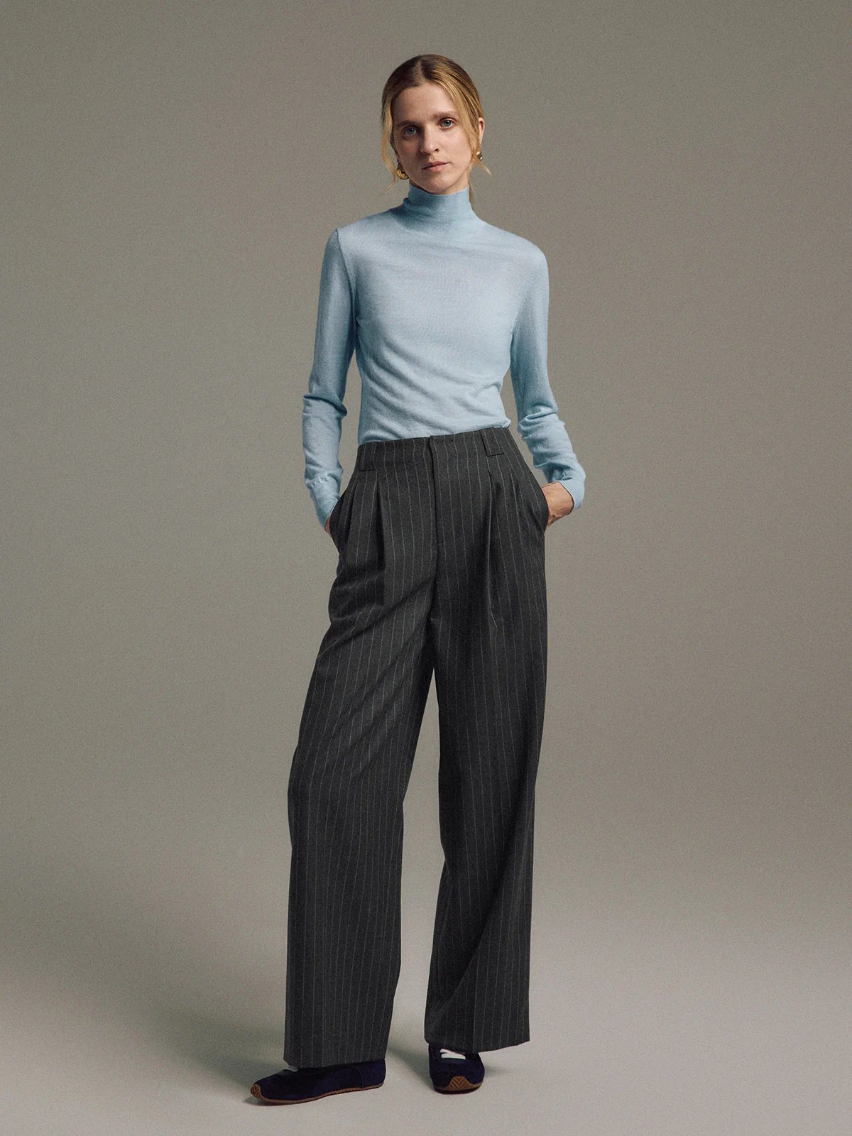 Side view of model wearing Dark Gray wool striped wide-leg trousers with hands in pockets and hair tied back.