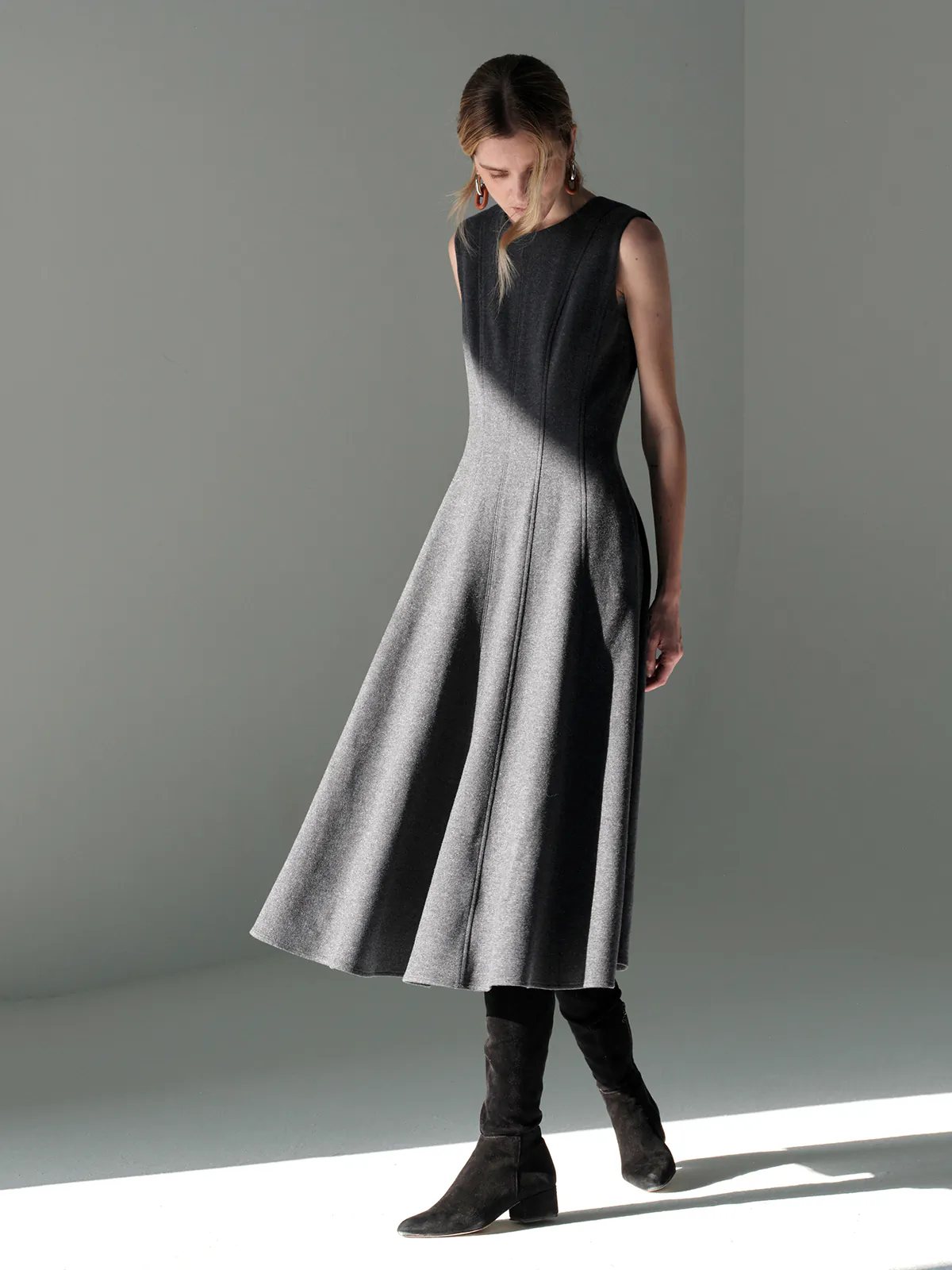 Front view of model in a sleeveless dark gray wool knit A-line dress layered over a light shirt, hands in pockets, with boots.