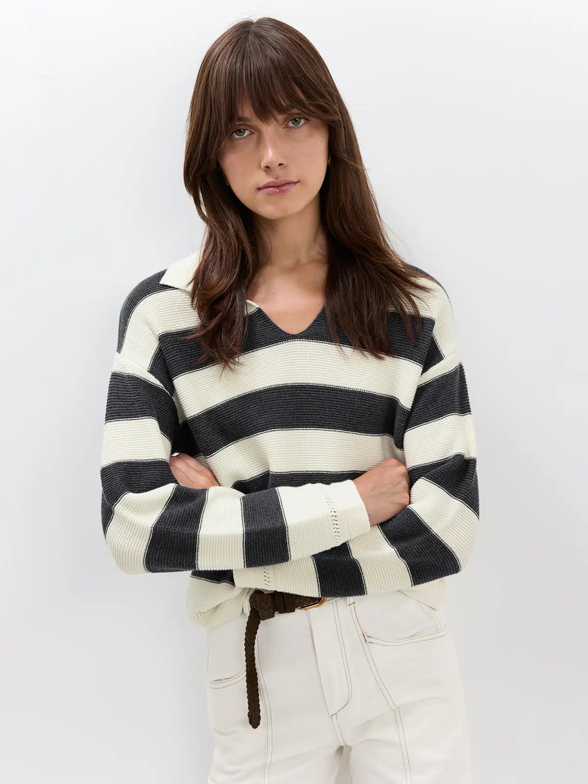 Full-front view of model in a Grey/White striped V-neck jumper tucked into white trousers with a belt and loafers.
