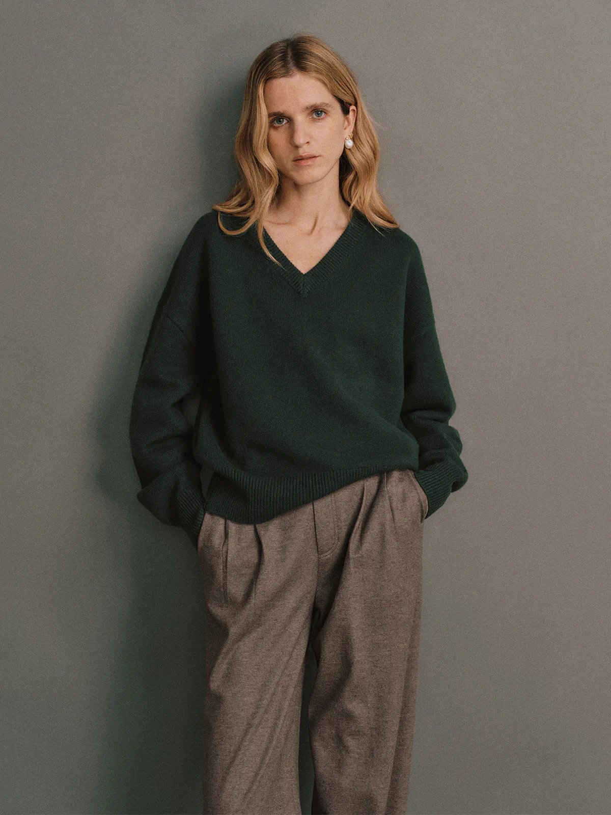 Side view of the Bottle Green CARRIE Merino Wool V-Neck Jumper worn with pleated trousers and hands in pockets.