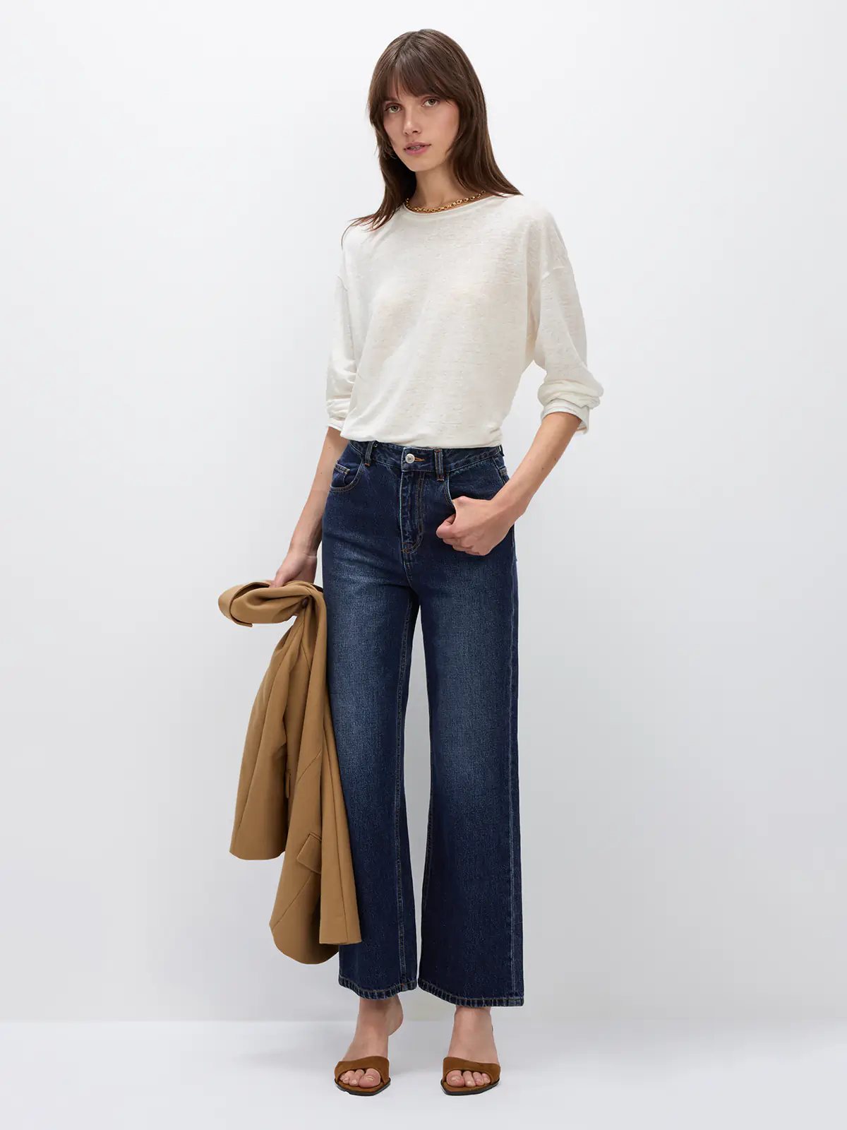 Side view of model in a white lightweight long-sleeve linen t-shirt with loose silhouette, holding a coat over dark wide-leg jeans.