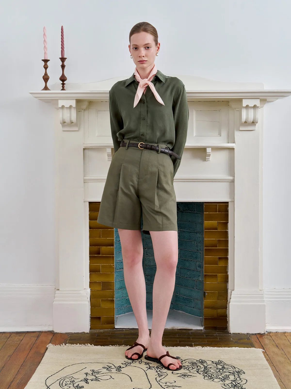 Model standing before fireplace wearing olive shirt, shorts, and blush pink TIANA scarf tied in bow