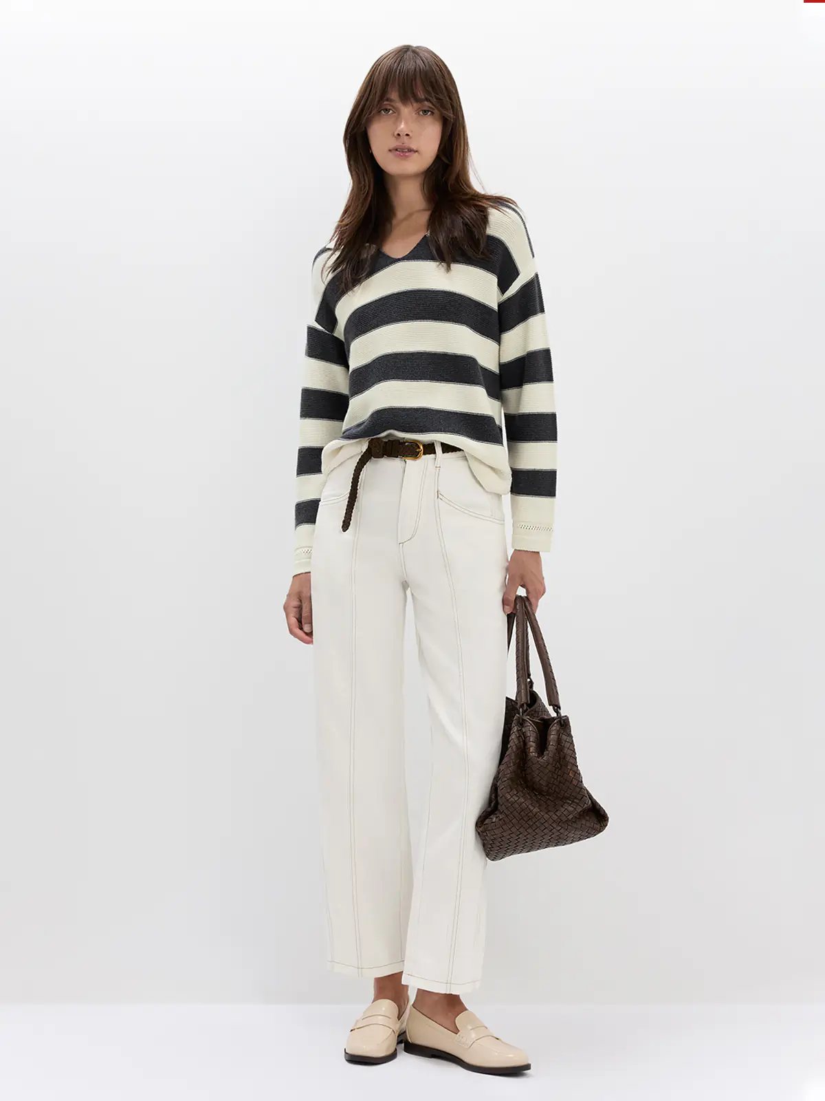Side view of model wearing a Grey/White striped V-neck jumper with ribbed sleeves, white trousers, and a belt.