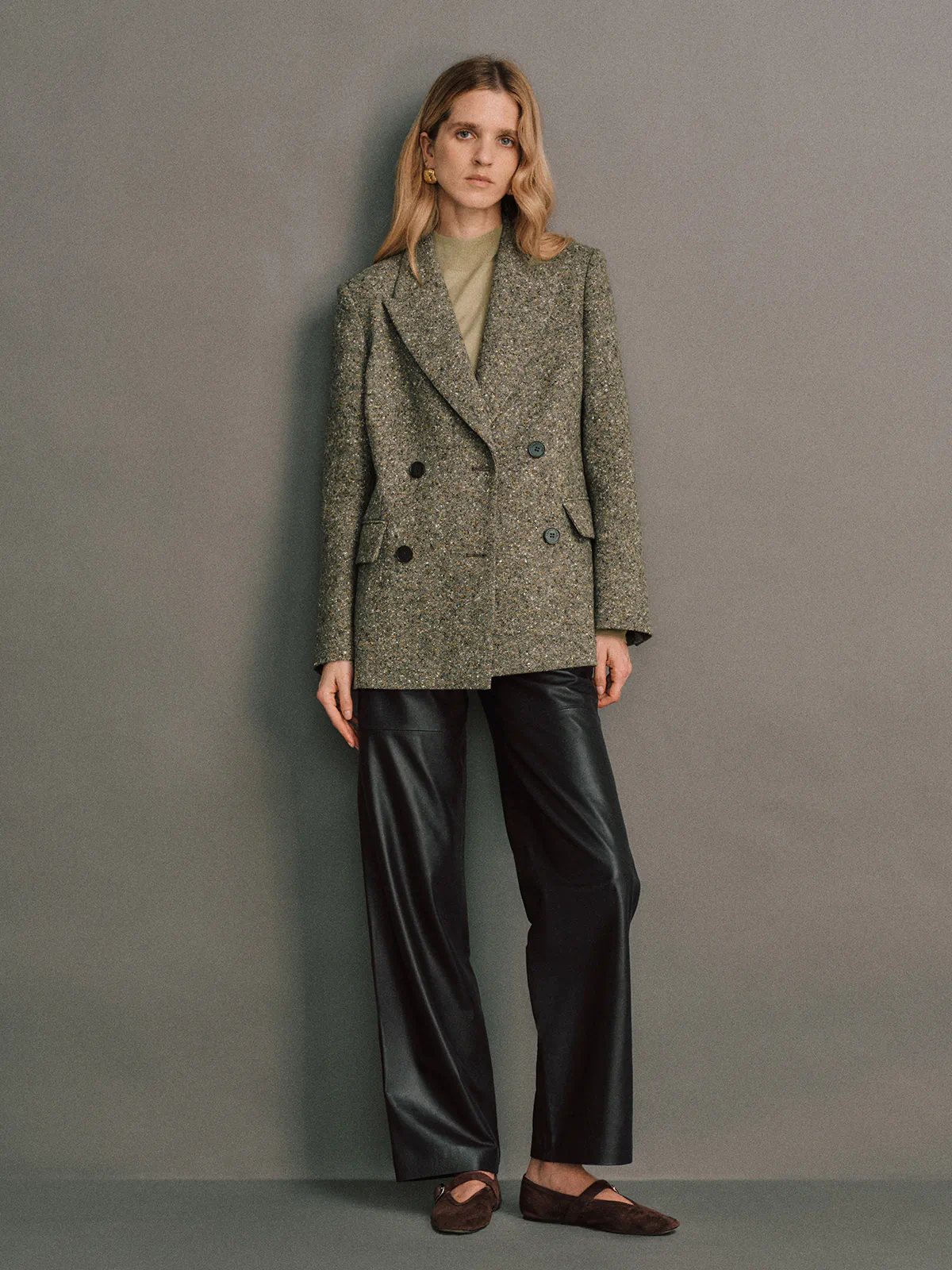 Full-length side view of a model in a green wool flecked tweed double-breasted blazer, wide-leg pants, sneakers, and a cap.
