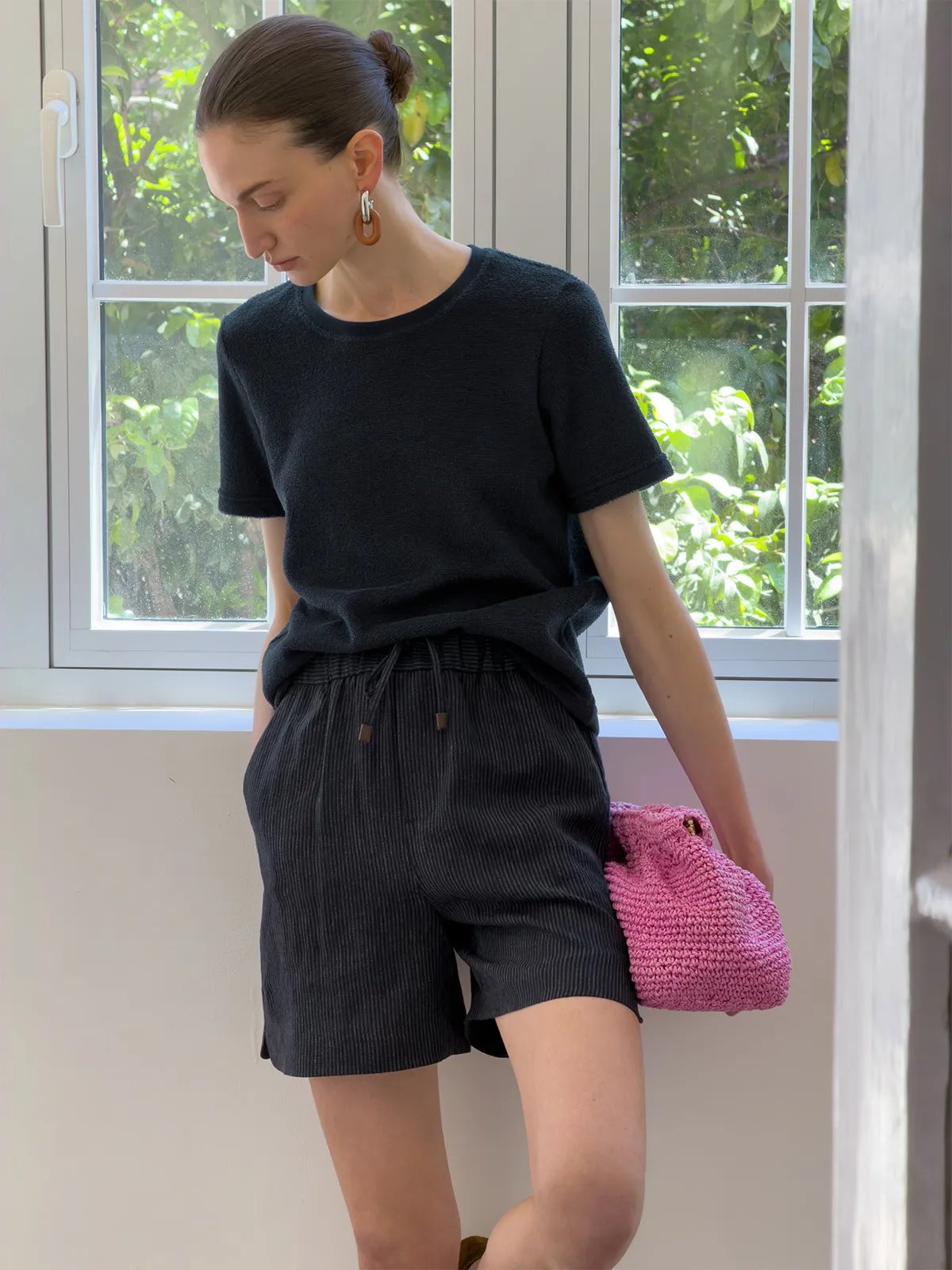 Model in black textured tee and NEVAEH shorts holding pink crocheted bag by window