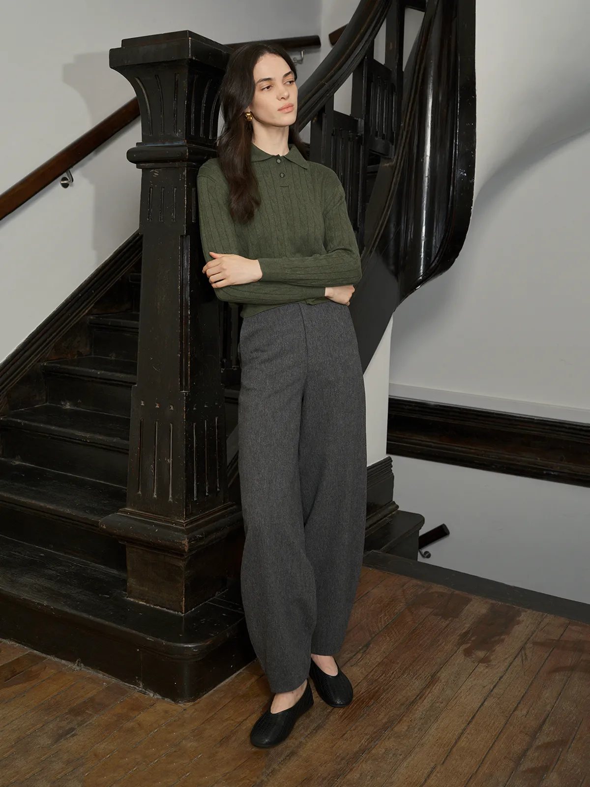 Model in olive green EMERSON Jumper with arms crossed, leaning against black staircase post—confident minimalist aesthetic
