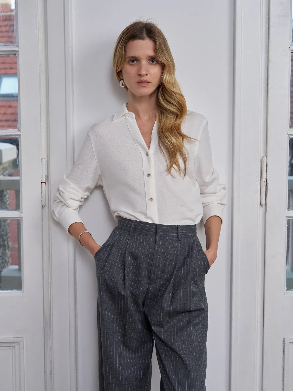 Front-facing model in ivory COLETTE pique shirt and grey pinstripe trousers, hands in pockets, minimalist interior setting
