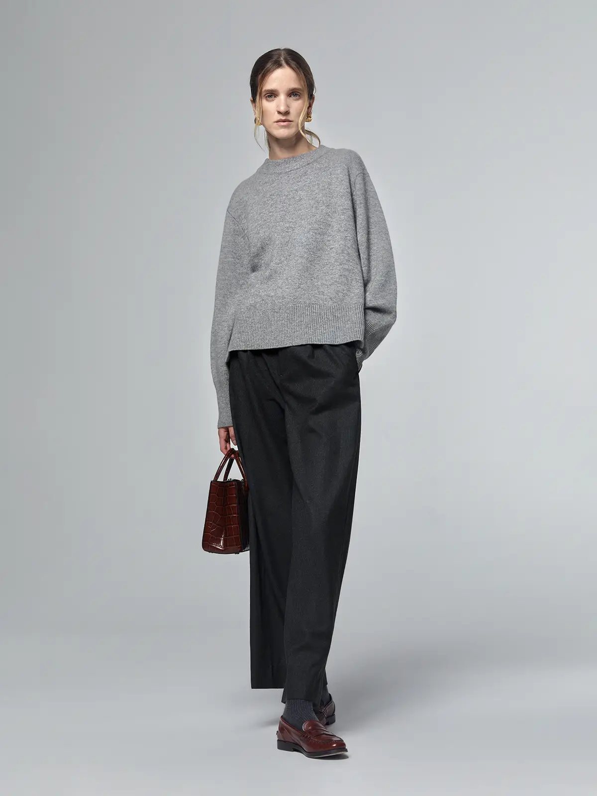 Model wearing a grey merino wool sweater with metal buttons and ribbed hem, paired with dark trousers and holding a bag.